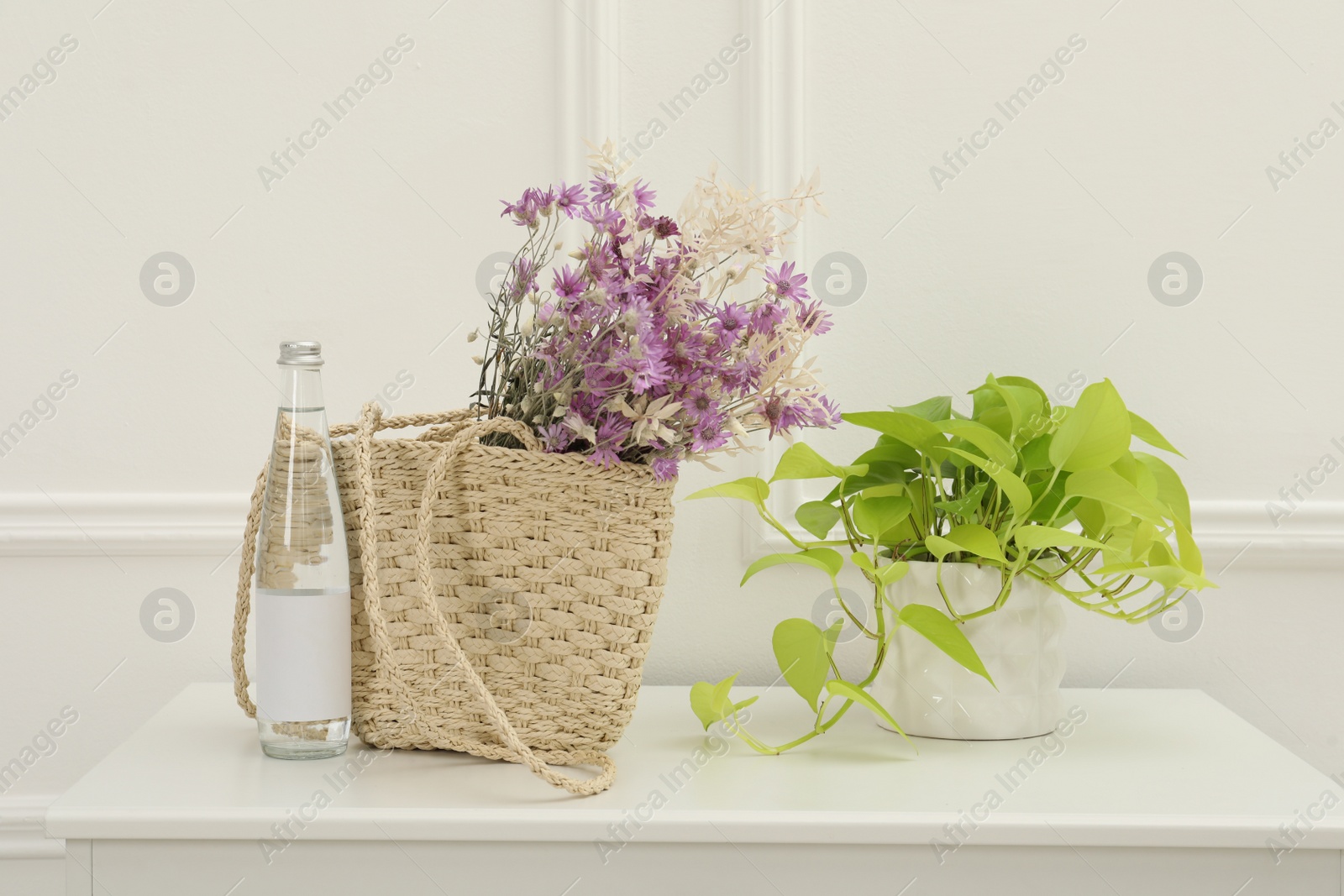 Photo of Stylish beach bag with beautiful wildflowers near bottle of water and houseplant on cabinet in room