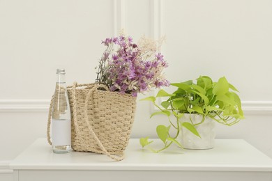 Stylish beach bag with beautiful wildflowers near bottle of water and houseplant on cabinet in room