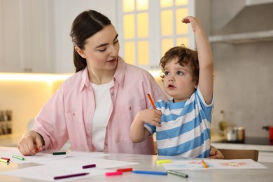 Mother and her little son drawing with colorful markers at table in kitchen