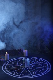 Photo of Natural stones for zodiac signs, drawn astrology chart and burning candles on dark blue table, space for text