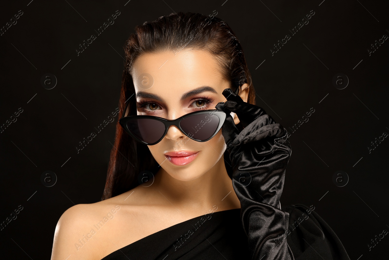 Photo of Portrait of beautiful young woman in sunglasses and elegant evening gloves on black background