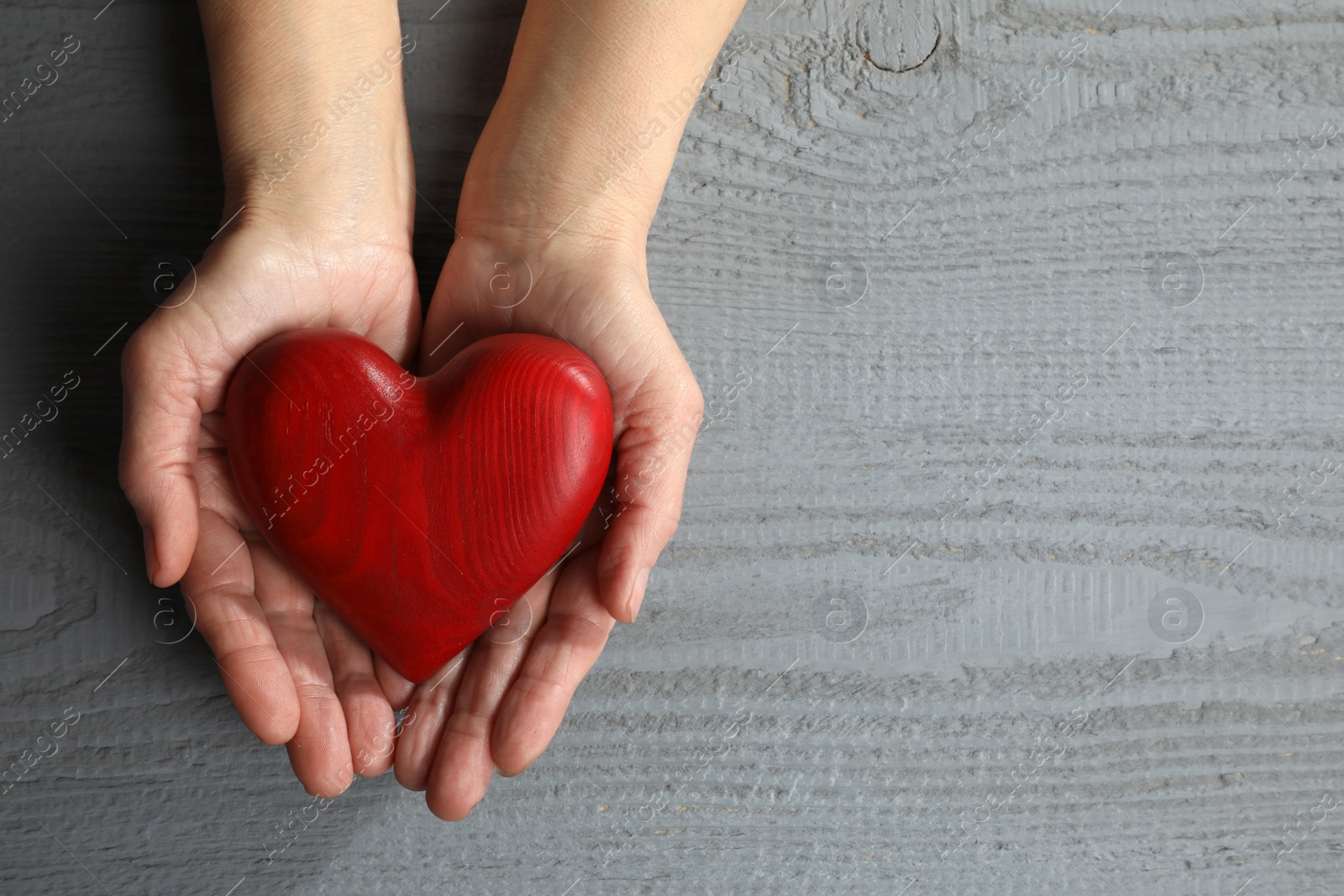 Photo of Elderly woman holding red heart in hands at grey wooden table, top view. Space for text