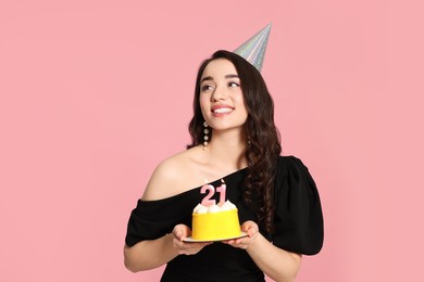 Coming of age party - 21st birthday. Smiling woman holding delicious cake with number shaped candles on pink background