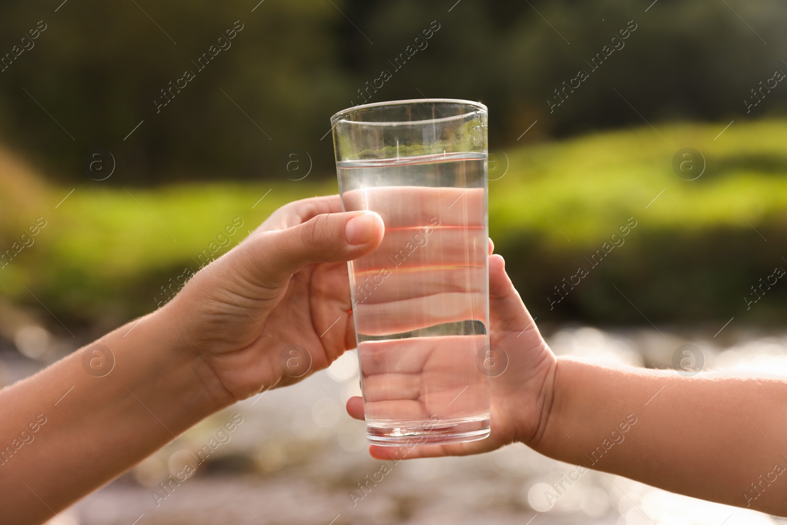 Photo of Mother giving her daughter glass of fresh water near stream on sunny day, closeup