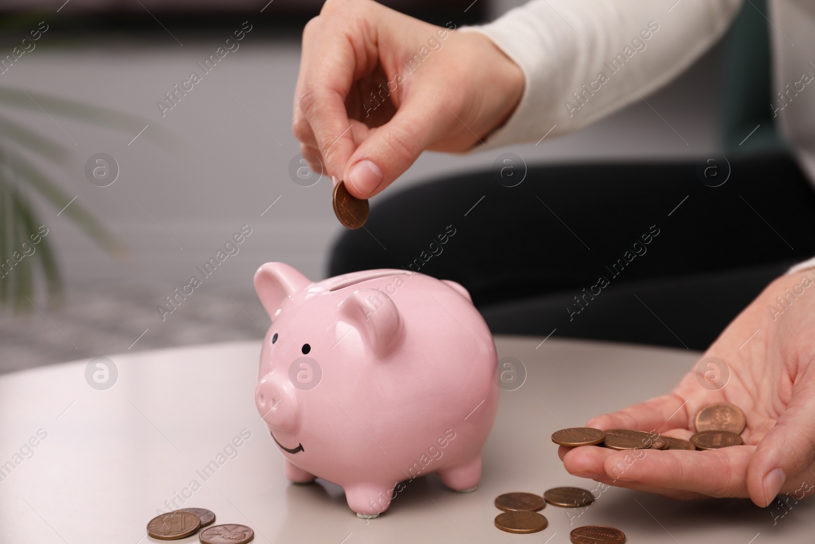 Photo of Woman putting coin into piggy bank at table indoors, closeup