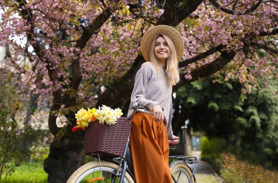 Beautiful young woman with bicycle and flowers in park on pleasant spring day