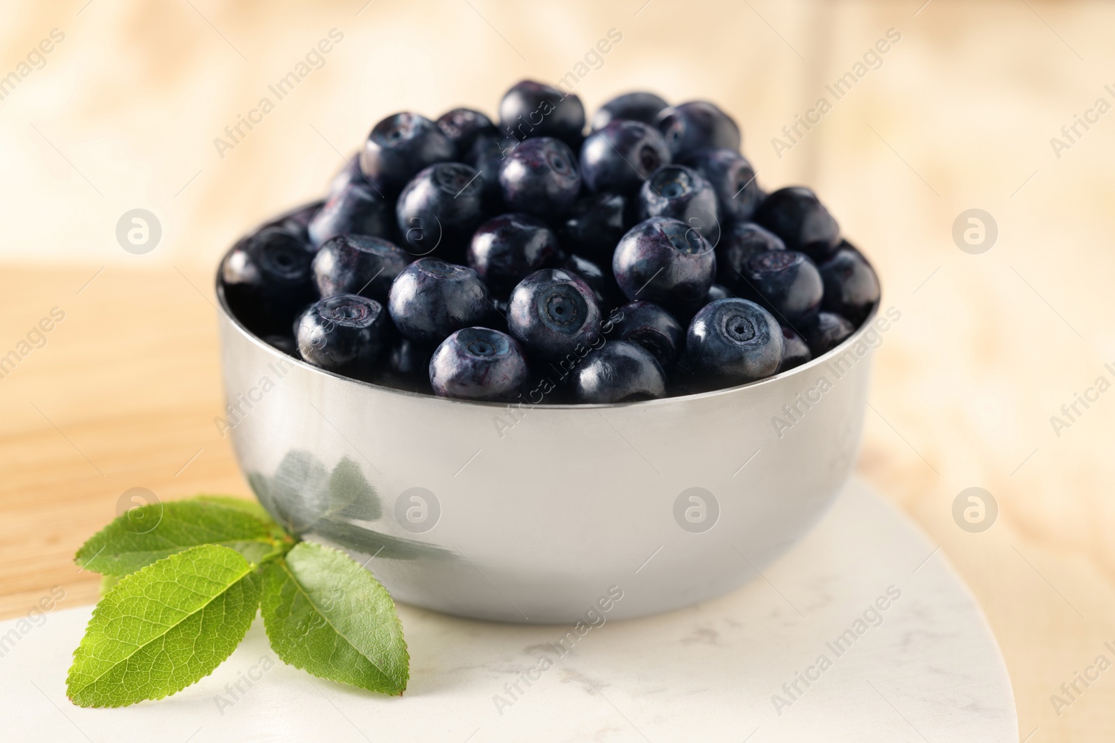 Photo of Bowl of tasty fresh bilberries and green leaves on light table, closeup