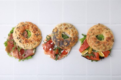 Tasty bagel sandwiches on white tiled table, flat lay