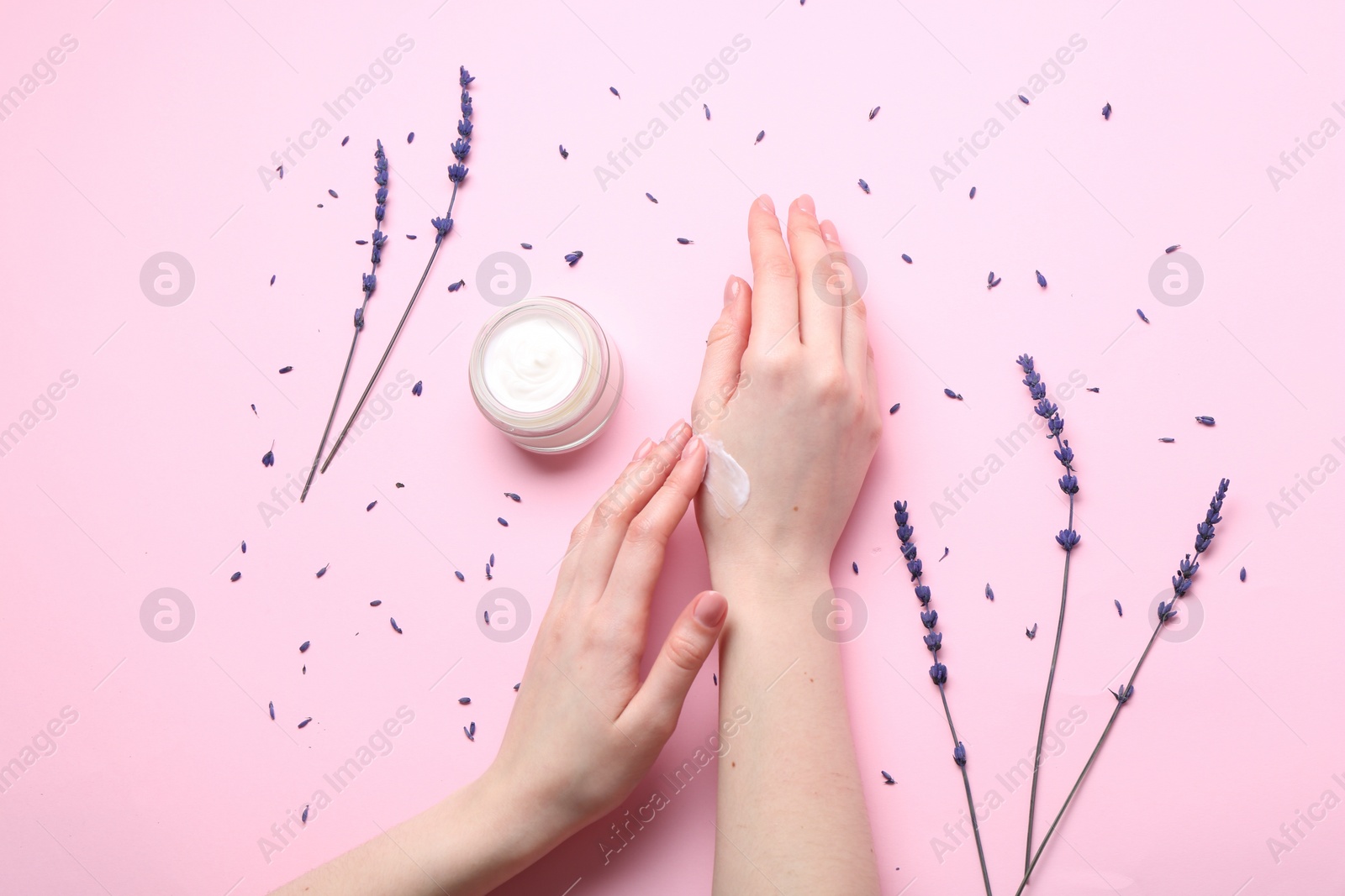 Photo of Woman applying hand cream and lavender flowers on pink background, top view