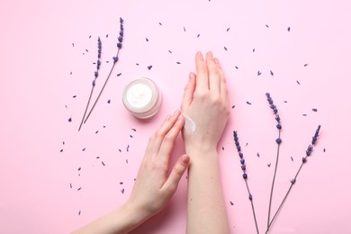 Photo of Woman applying hand cream and lavender flowers on pink background, top view