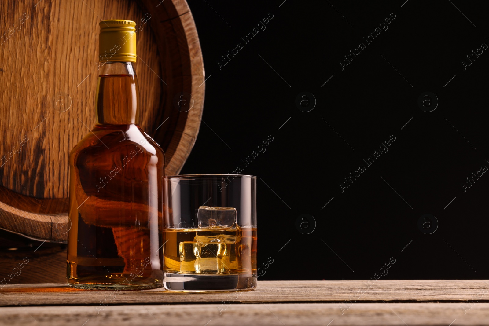 Photo of Whiskey with ice cubes in glass, bottle and barrel on wooden table against black background, space for text