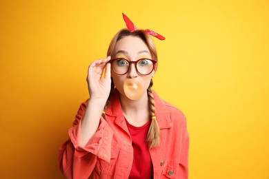 Photo of Fashionable young woman with braids and bright makeup blowing bubblegum on yellow background