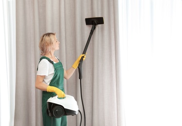 Photo of Female janitor removing dust from curtain with steam cleaner indoors