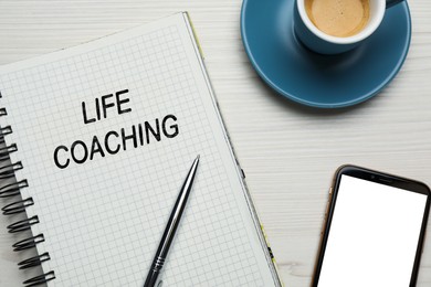 Phrase Life Coaching written in notebook, pen, smartphone and cup coffee on white wooden table, flat lay