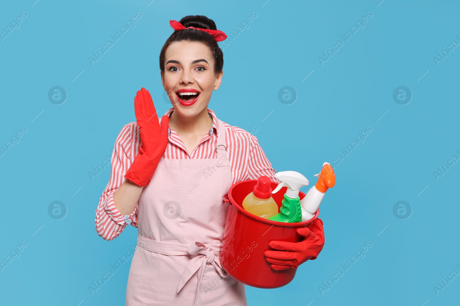 Photo of Emotional housewife holding bucket with cleaning supplies on light blue background