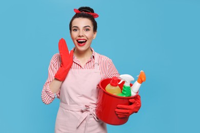 Photo of Emotional housewife holding bucket with cleaning supplies on light blue background