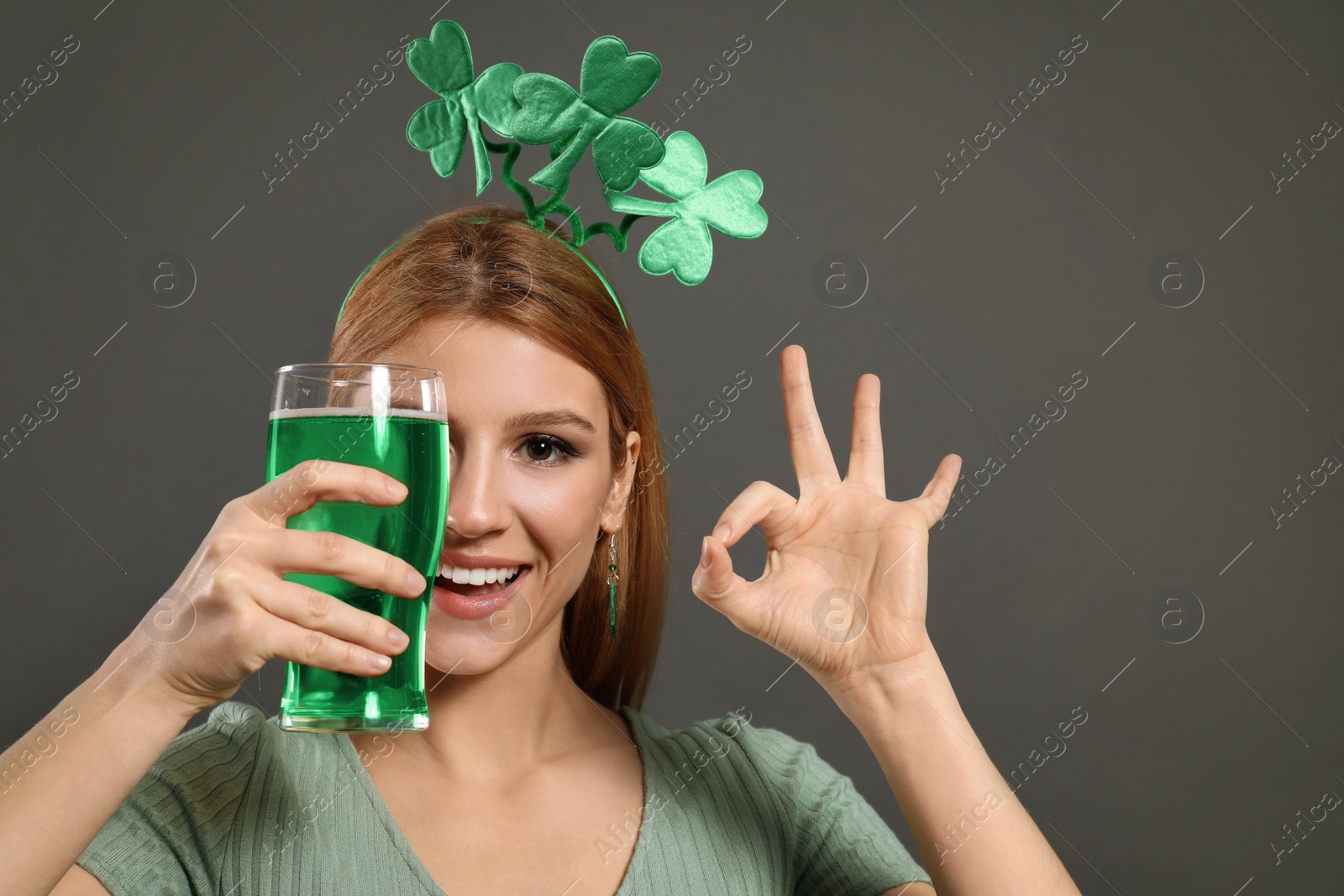 Photo of Young woman with clover headband and green beer on grey background. St. Patrick's Day celebration