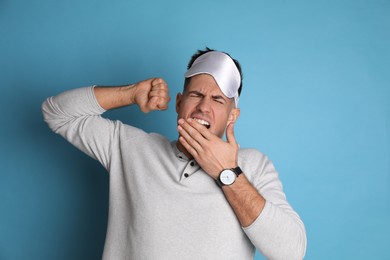 Tired man with sleeping mask yawning on light blue background