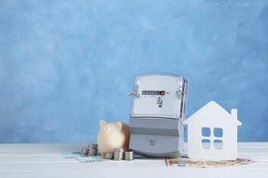 Photo of Electricity meter, piggy bank, house model and money on white wooden table. Space for text