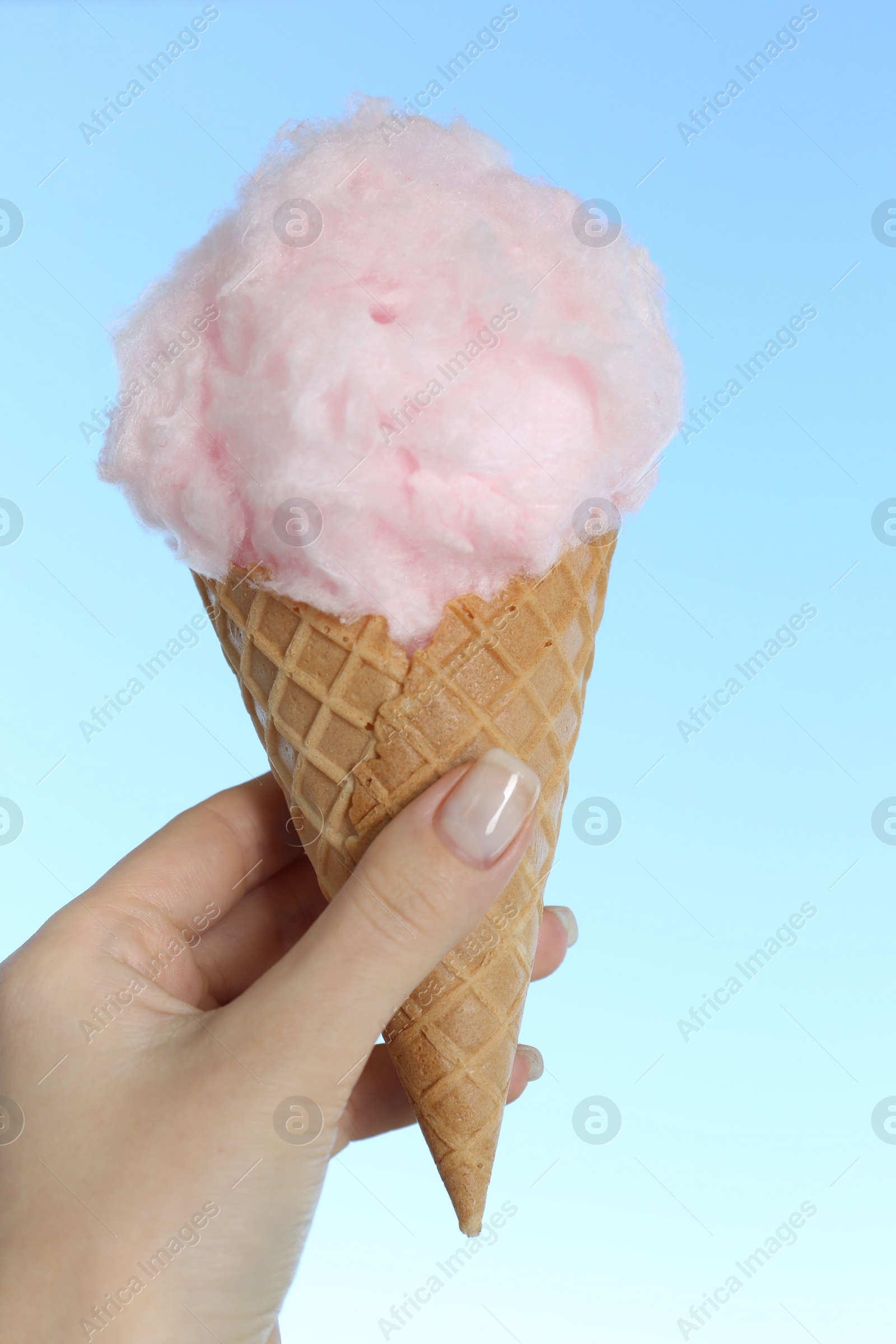 Photo of Woman holding waffle cone with sweet cotton candy on light blue background, closeup