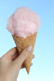 Woman holding waffle cone with sweet cotton candy on light blue background, closeup