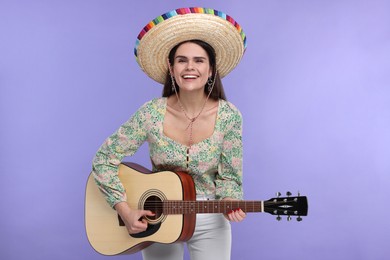 Young woman in Mexican sombrero hat playing guitar on violet background