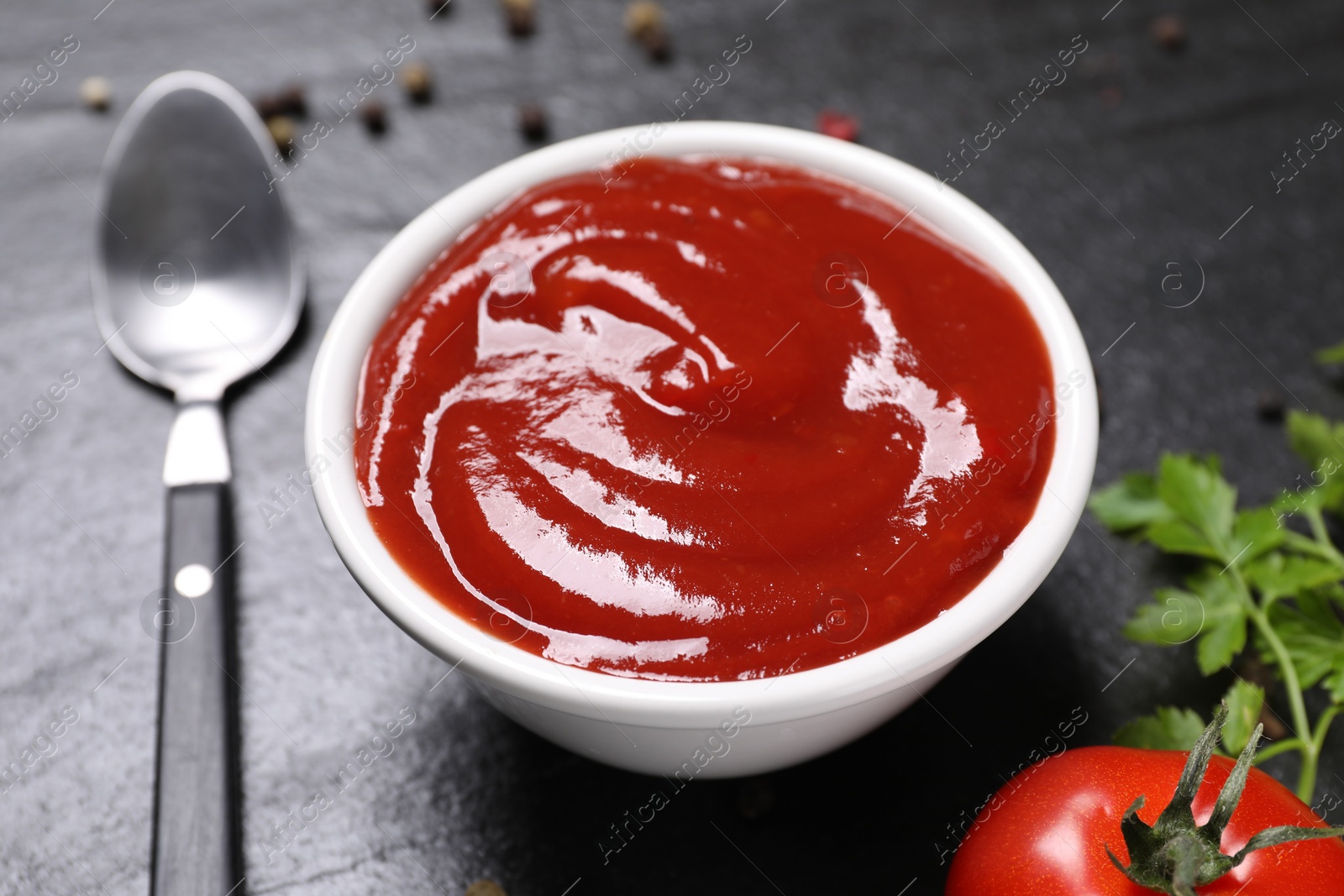 Photo of Organic ketchup in bowl, fresh tomato and spoon on black table, closeup. Tomato sauce