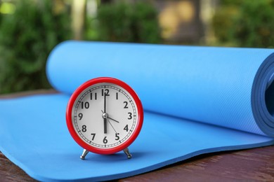Photo of Alarm clock and fitness mat on wooden table outdoors. Morning exercise