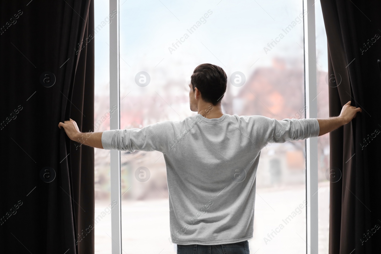 Photo of Man opening window curtains at home, back view