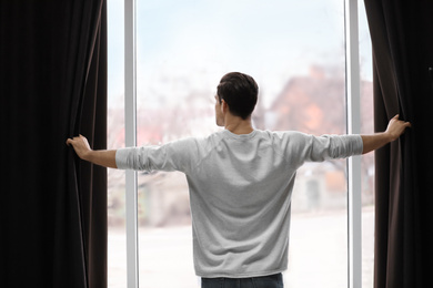 Photo of Man opening window curtains at home, back view