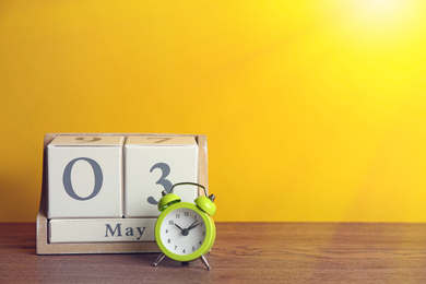 Wooden block calendar and alarm clock on table against yellow background. Space for text