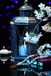 Photo of Arabic lantern, Quran, misbaha, burning candle and flowers on mirror surface against blurred lights at night