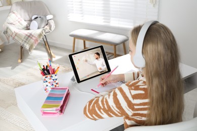 Image of E-learning. Little girl taking notes during online lesson at table indoors
