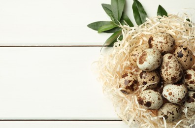 Photo of Nest with many speckled quail eggs and green leaves on white wooden table. Space for text