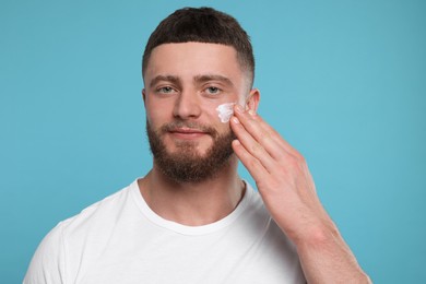 Handsome man applying cream onto his face on light blue background