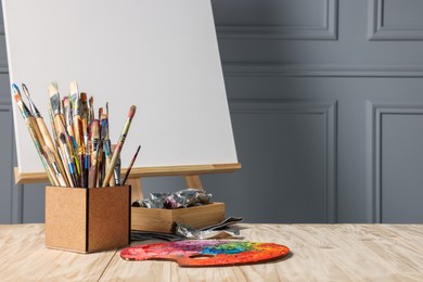 Photo of Easel with blank canvas and different art supplies on wooden table near grey wall. Space for text