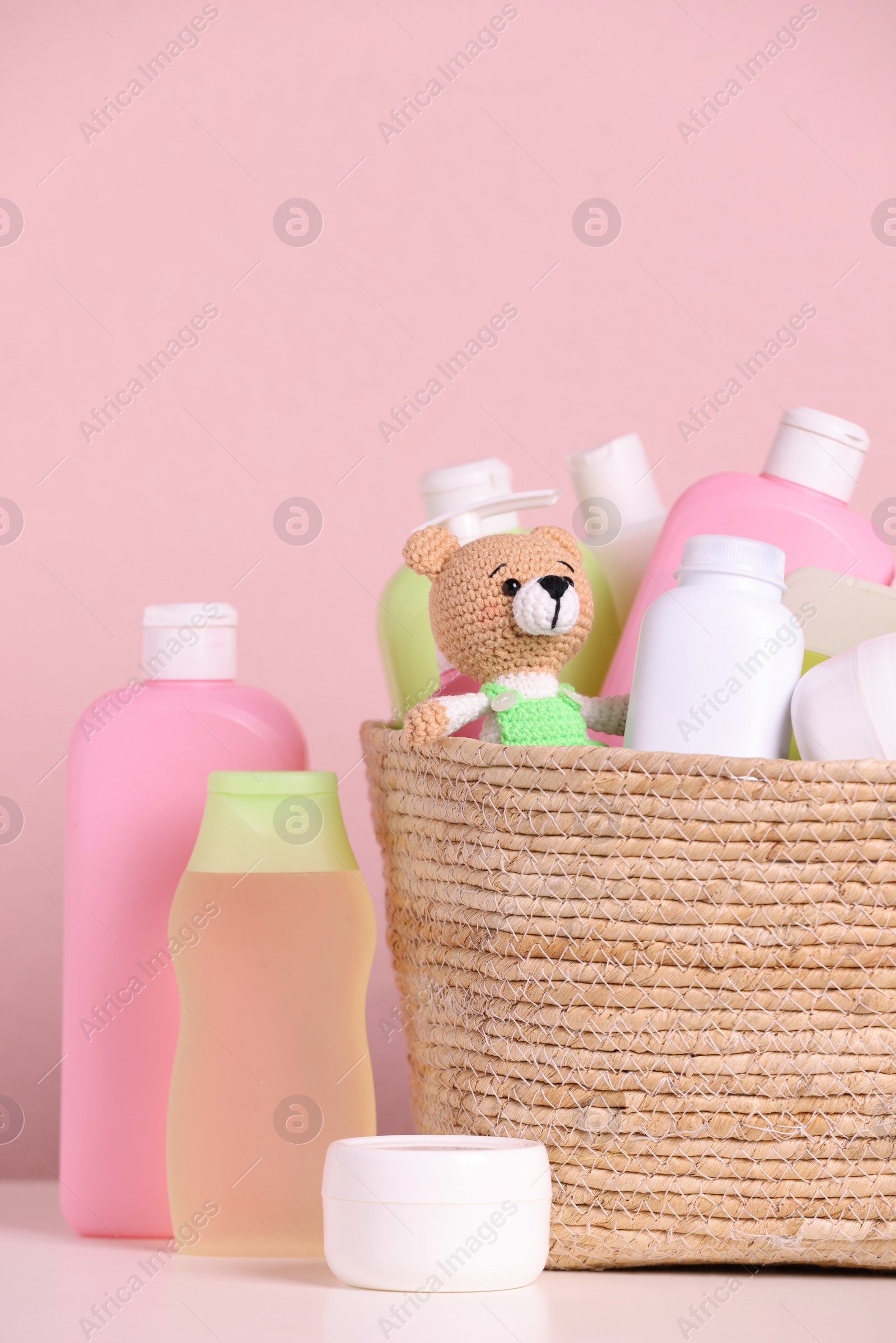 Photo of Baby cosmetic products, bath accessories and toy in wicker basket on white table against pink background