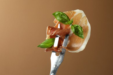 Photo of Fork with slice of tasty jamon and basil leaves on brown background, closeup