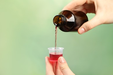 Photo of Woman pouring cough syrup into measuring cup on light green background, closeup