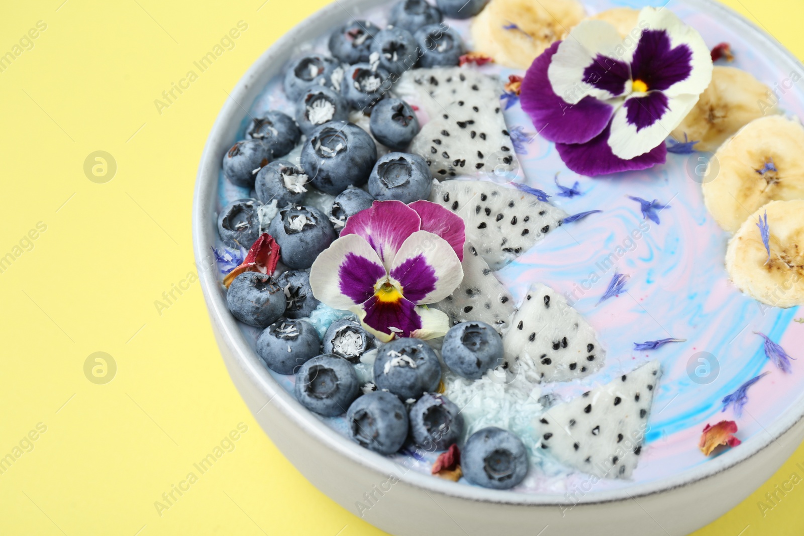 Photo of Delicious smoothie bowl with fresh fruits, blueberries and flowers on yellow background, closeup