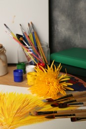 Photo of Canvas with colorful painting, yellow chrysanthemum and brushes on table