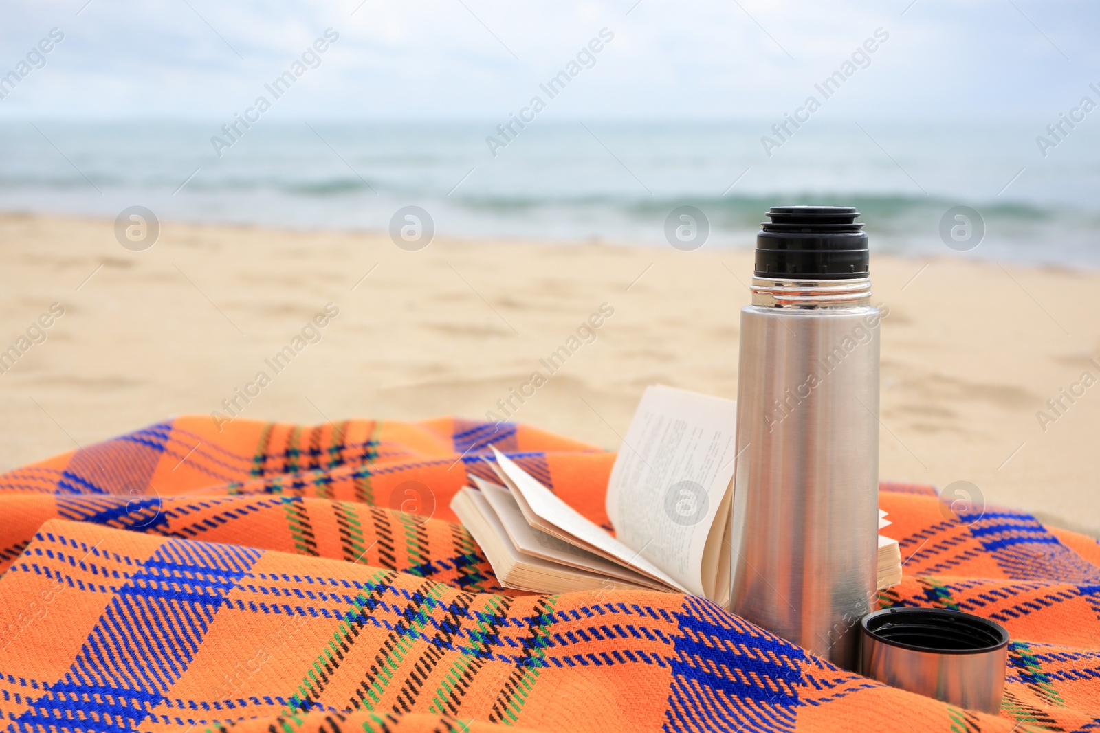 Photo of Metallic thermos with hot drink, open book and plaid on sandy beach near sea, space for text