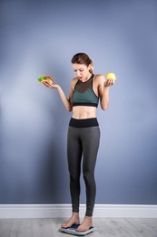 Photo of Woman holding tasty sandwich and apple while measuring her weight on floor scales near color wall. Weight loss motivation