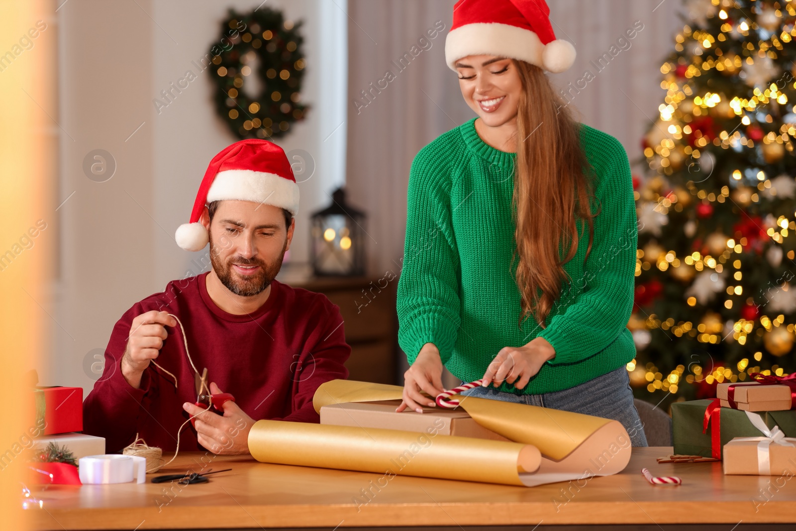 Photo of Happy couple in Santa hats decorating Christmas gift at table in room
