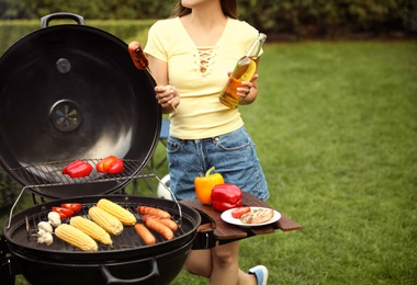 Photo of Woman with beer and sausage near barbecue grill outdoors, closeup