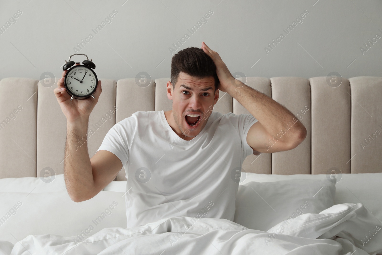 Photo of Emotional man with alarm clock in bed. Being late because of oversleeping