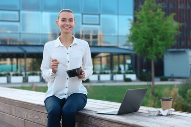 Photo of Smiling businesswoman with lunch box sitting near laptop and paper cup outdoors