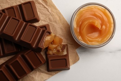 Tasty chocolate bars and bowl of caramel on white table, flat lay