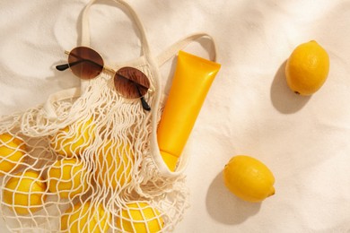 Photo of String bag with sunglasses, lemons and sunscreen on sand, flat lay