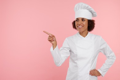 Photo of Happy female chef in uniform pointing at something on pink background. Space for text